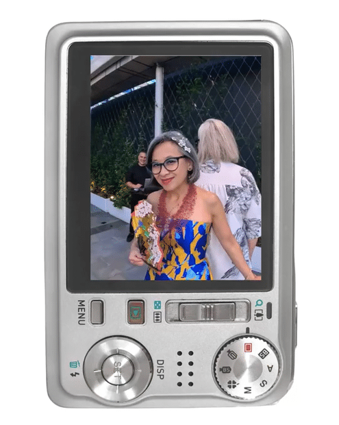 Digital camera screen showing Heather Wallace and Lan Tran co-founders of Be Savvy, with glasses, gray hair, and a floral dress holding a bouquet, standing outdoors at a social event.
