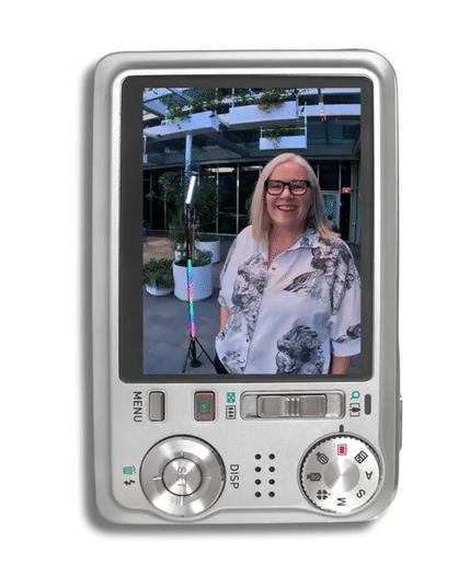 Heather Wallace co-founders of Be Savvy with blonde hair and glasses smiling inside a camera screen, wearing a white shirt with a black and gray floral pattern, outdoors near modern buildings.