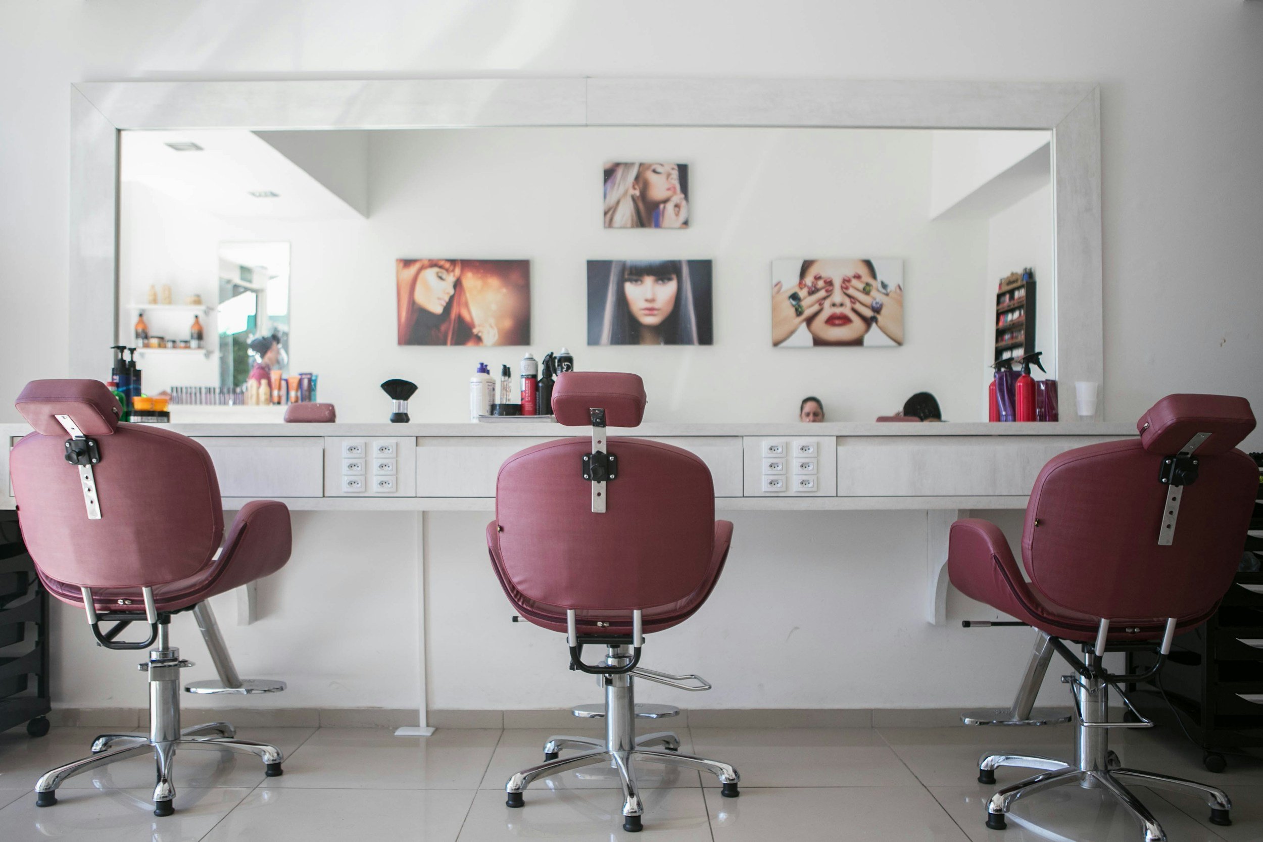 A salon station with three pink chairs facing a large mirror, salon supplies on the counter, and framed photos of women with styled hair on the wall.