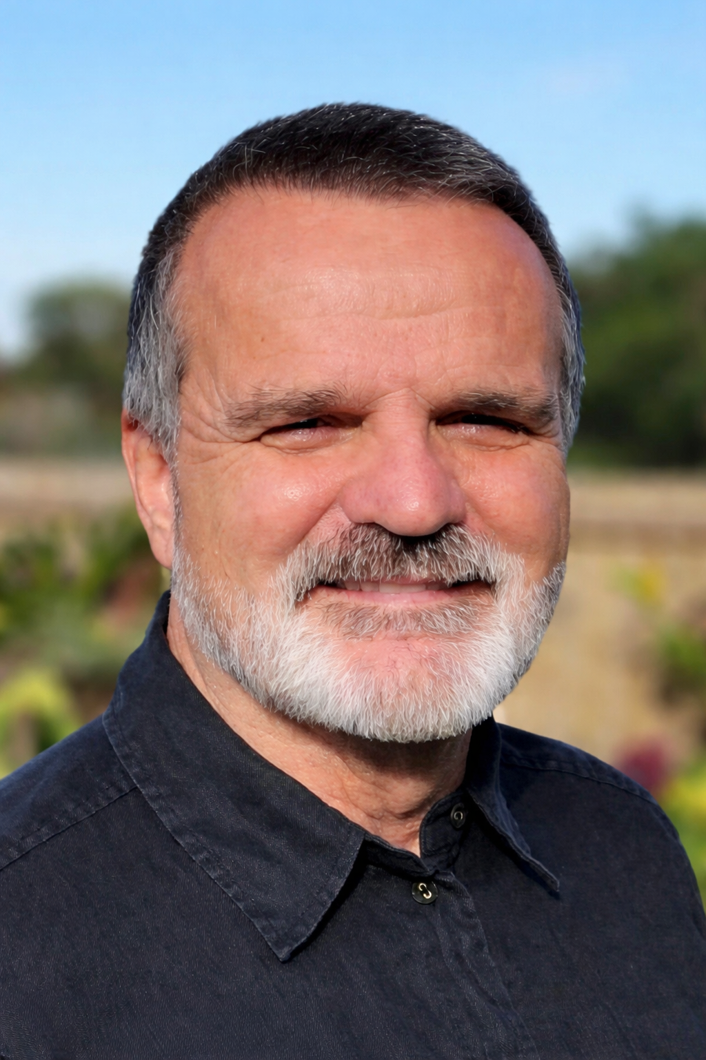 A middle-aged man with gray hair, a beard, and a mustache, smiling outdoors with a blurred background of greenery and blue sky.