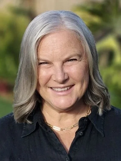 A smiling woman with shoulder-length gray hair, wearing a black shirt and a gold necklace, outdoors with blurred green foliage in the background.