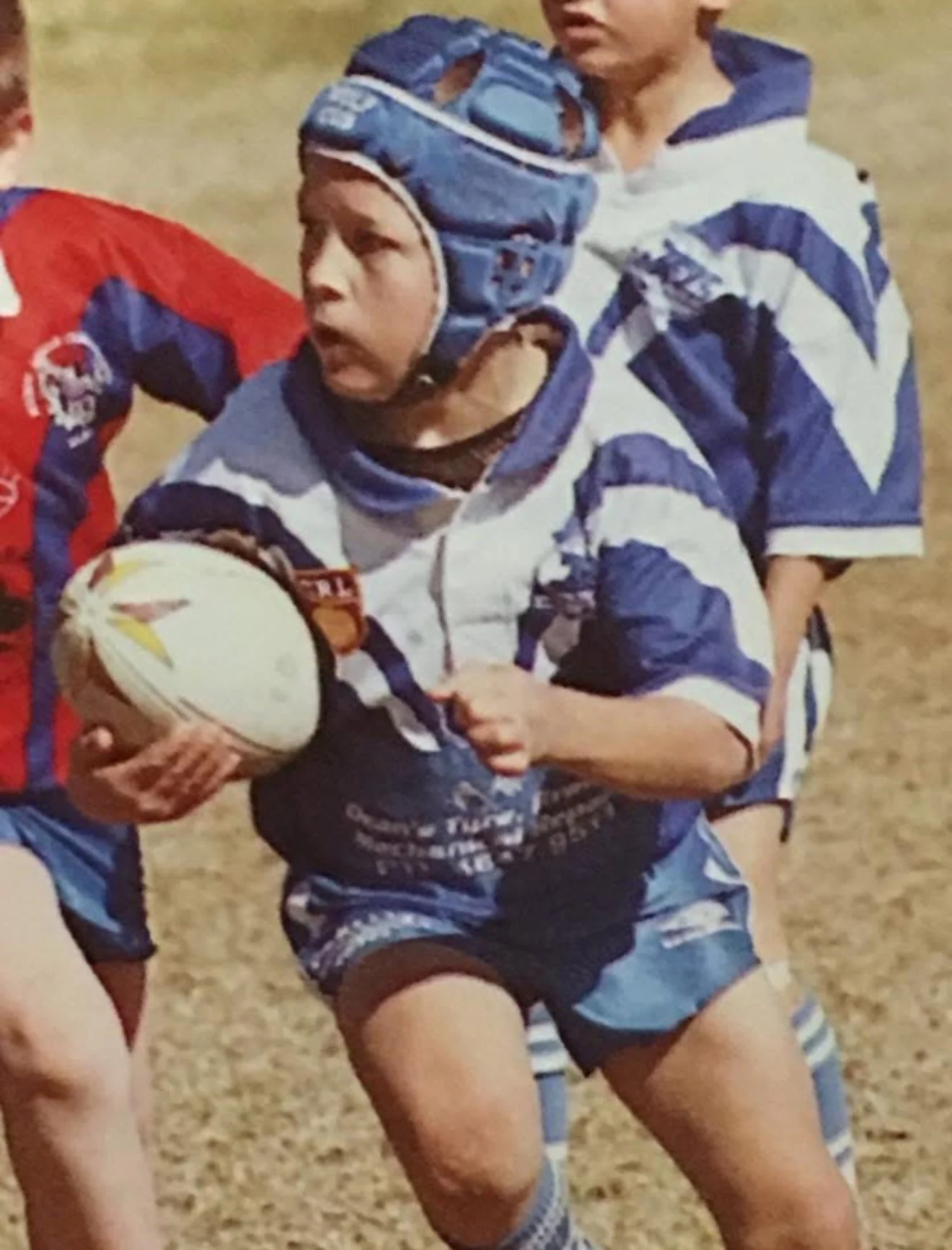 A young rugby player wearing a blue and white striped jersey, blue helmet, and blue shorts, holding a rugby ball, running on a grassy field during a game.