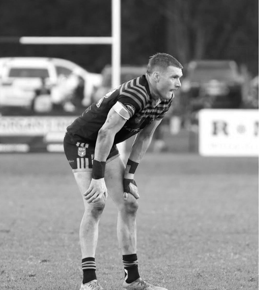 A rugby player in a striped uniform bending over on the field during a game, with parked cars and advertising signs in the background.