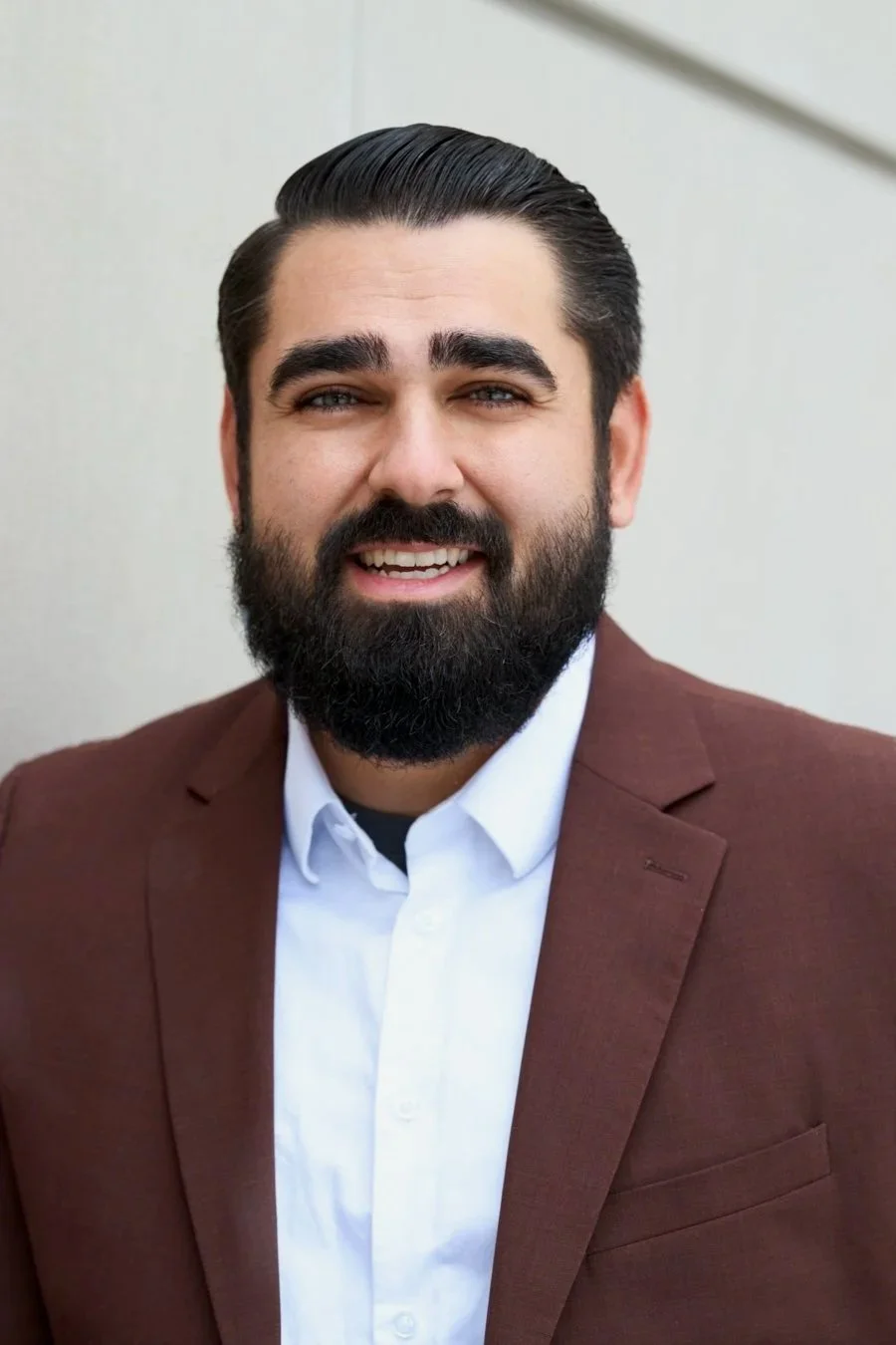 A smiling man with dark hair and a full beard, wearing a white shirt and a brown blazer, standing outdoors against a neutral background.