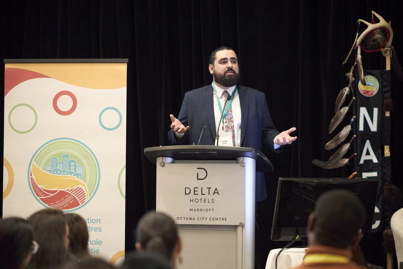 A man in a dark suit and tie is speaking at a podium during a conference at Delta Hotels by Marriott, Ottawa City Centre. Behind him are banners with colorful designs and symbols representing Indigenous culture.