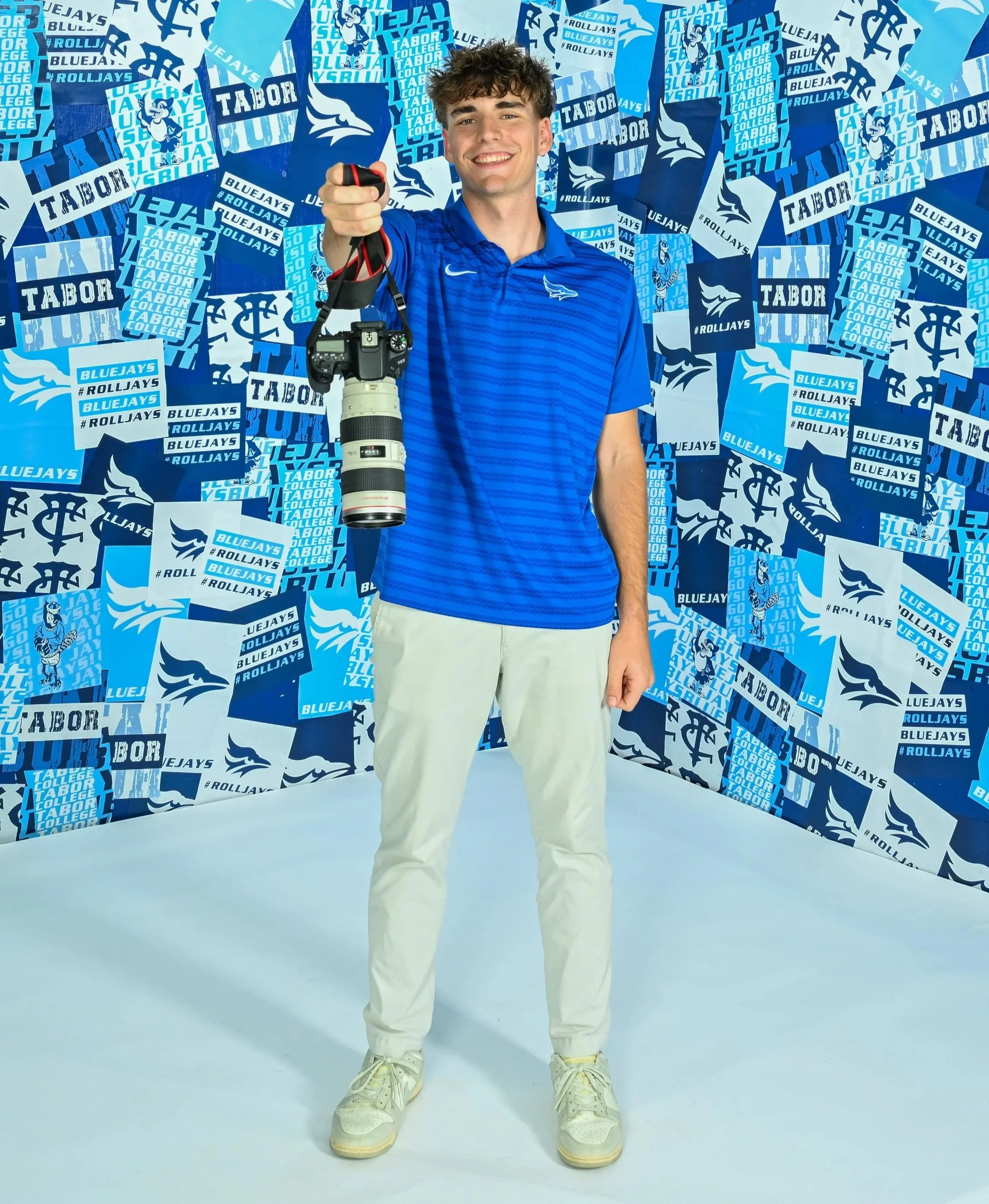 A young man is smiling and holding a camera with a telephoto lens in a photo studio with a blue-themed backdrop. The backdrop is decorated with various blue and white paper posters and banners displaying the words "BlueJays," "#RollJays," "Tabor College," and additional related graphics and team logos.