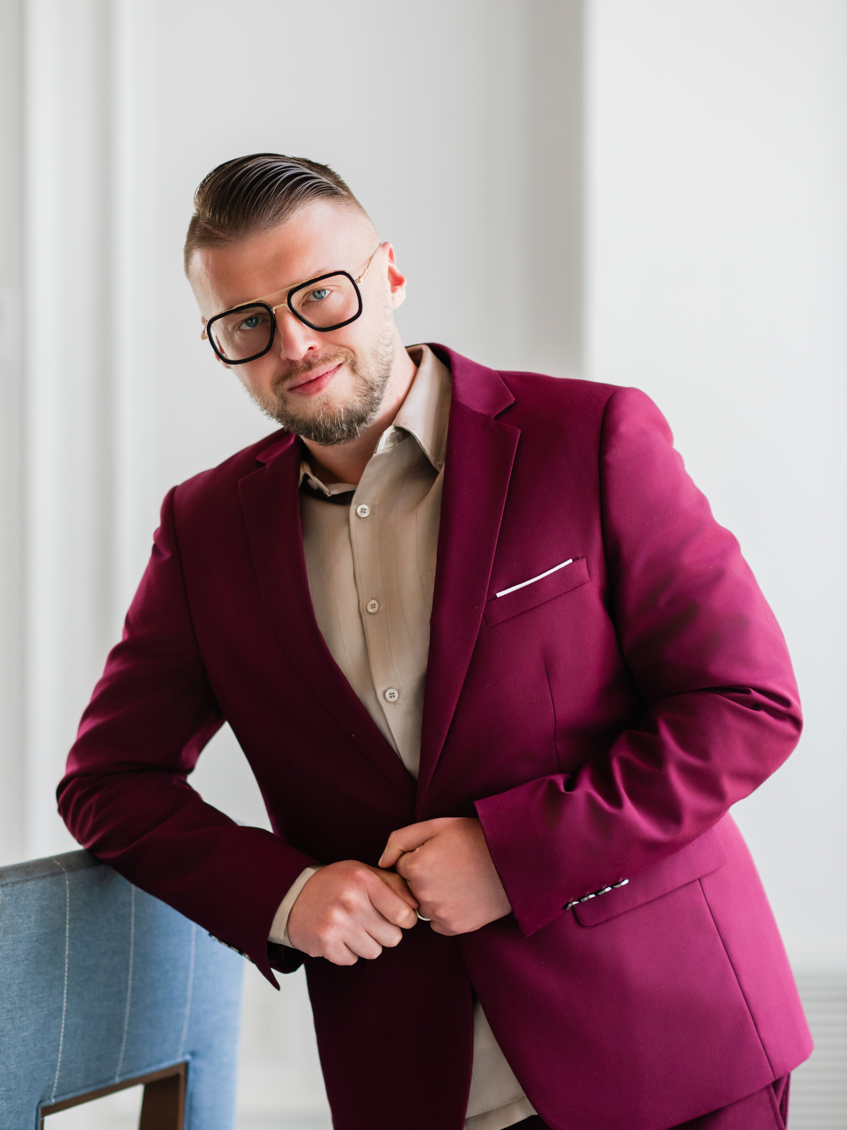 A man wearing a maroon blazer, beige shirt, black glasses, and sporting a beard, standing indoors with a neutral background.