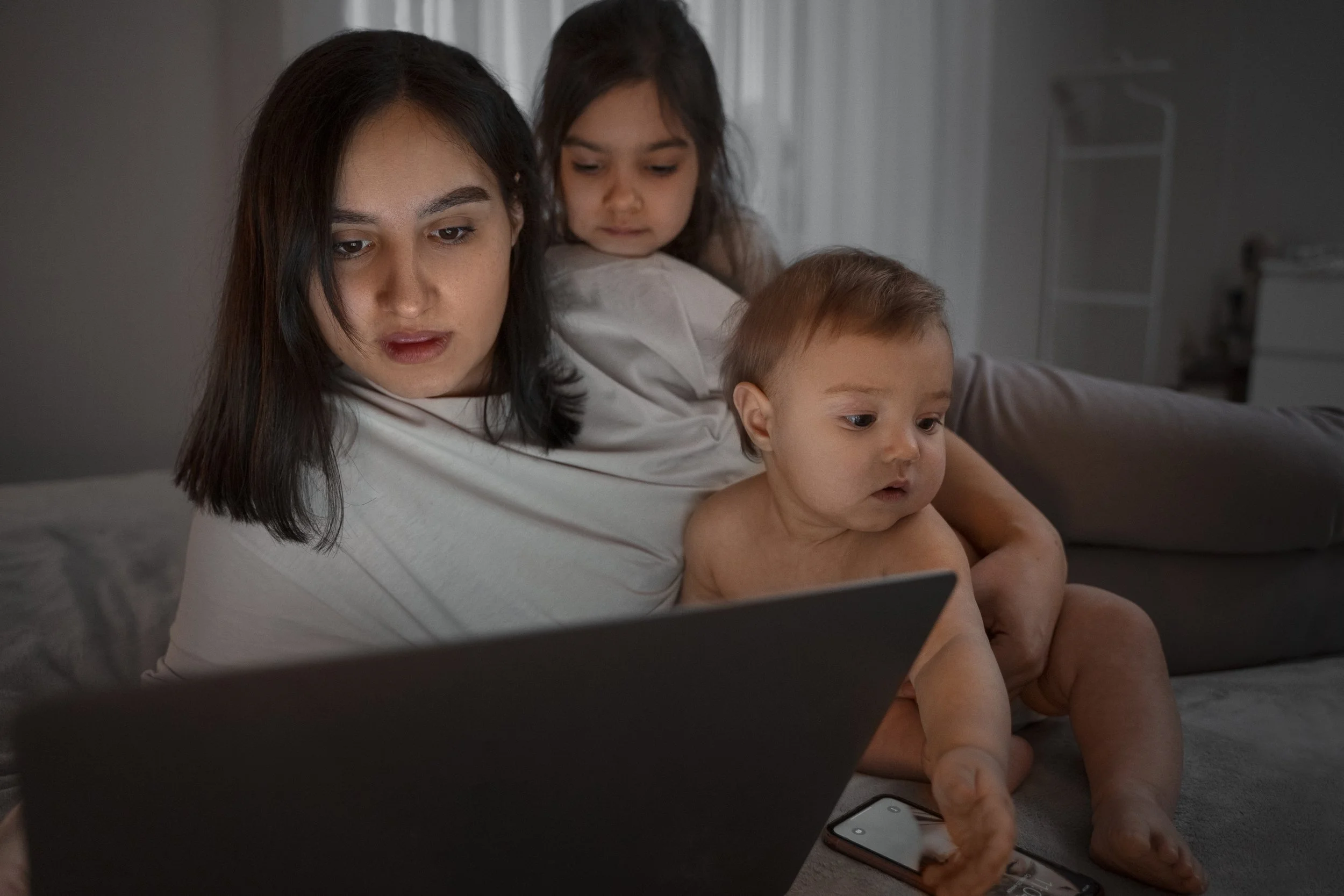 A woman and two children sitting on a bed, the woman is researching on a laptop in a dimly lit room. She has a baby on her lap and a toddler looking over her shoulder.