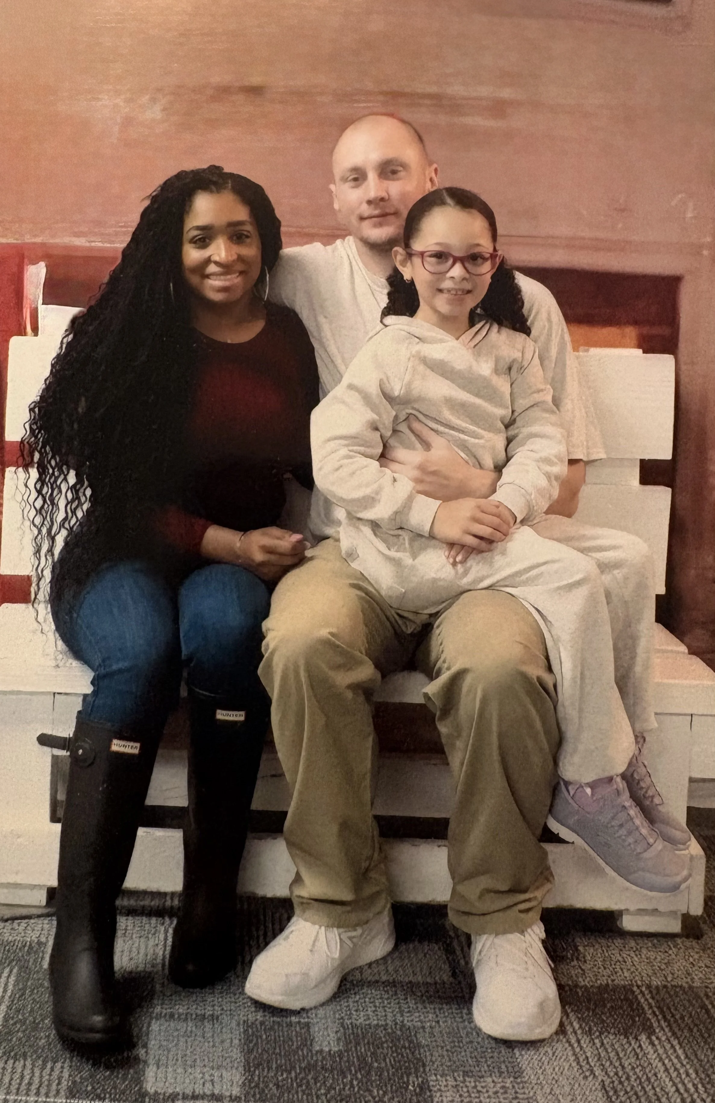 Chloe Appleberry and her family of three siton a wooden bench with Chloe on the left, her husband Tristan in the middle, and a their darling daughter with glasses sitting on his lap.