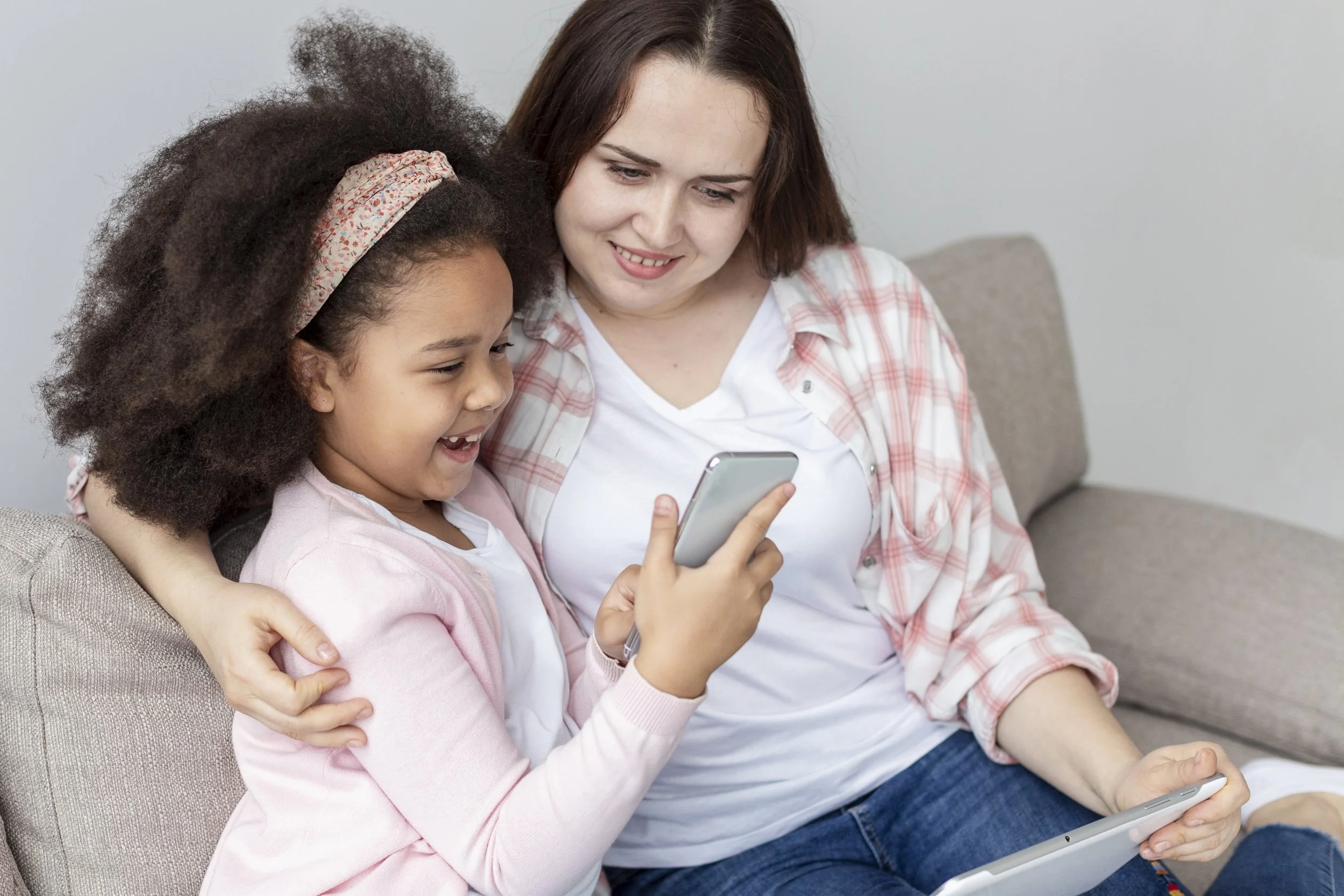 A woman and a young girl sitting on a beige couch, looking at a smartphone together, smiling and laughing. The woman has long dark hair. The happy child has thick curly hair pulled back in a pink headband and a bright smile.