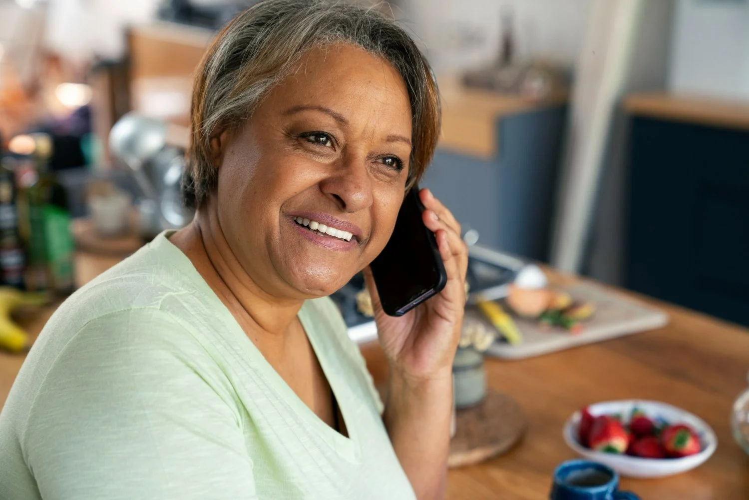 A middle-aged woman with short gray hair, smiling while talking on a mobile phone inside a kitchen. The background shows a kitchen counter with various items including strawberries and a bowl of strawberries, a cutting board, and other kitchen utensils.