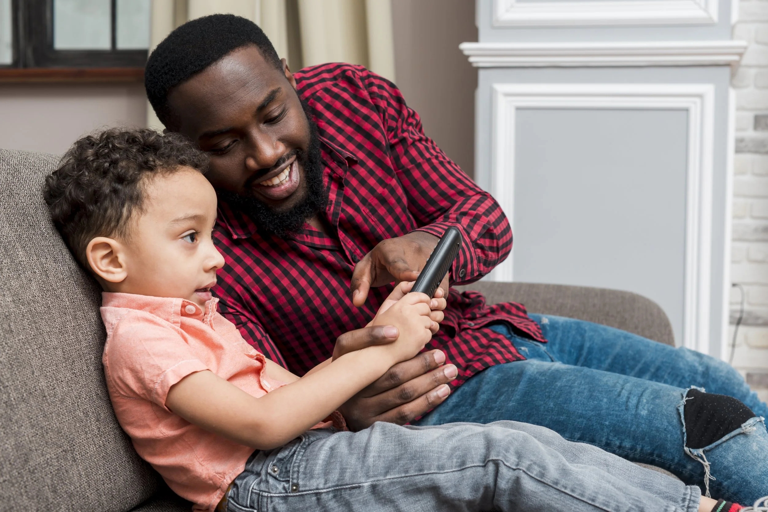A father and son sitting on a gray couch, engaged in a speakerphone call, in a cozy home setting.