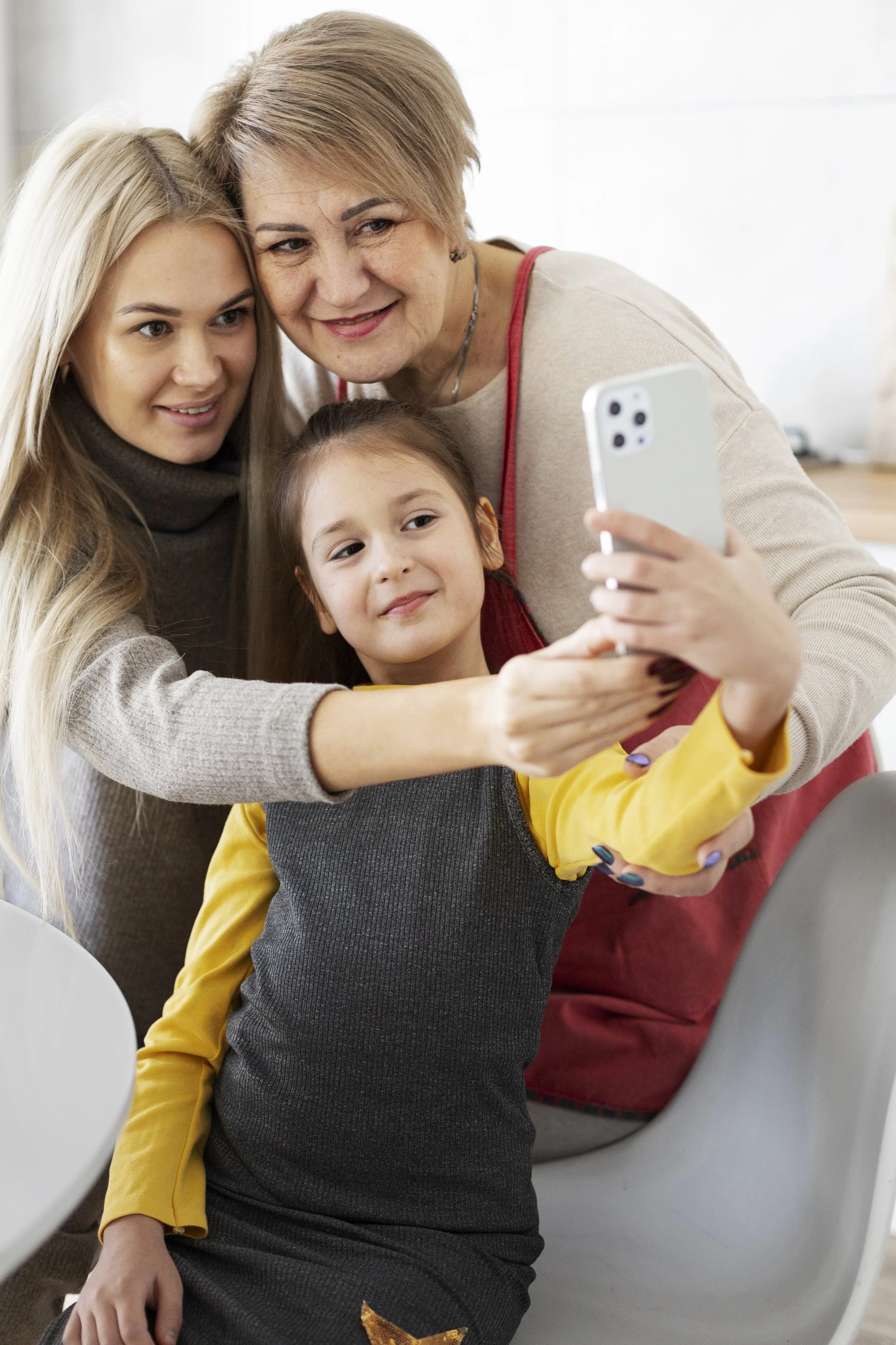 Three women and a young huddle to facetime with someone on a cell phone indoors.