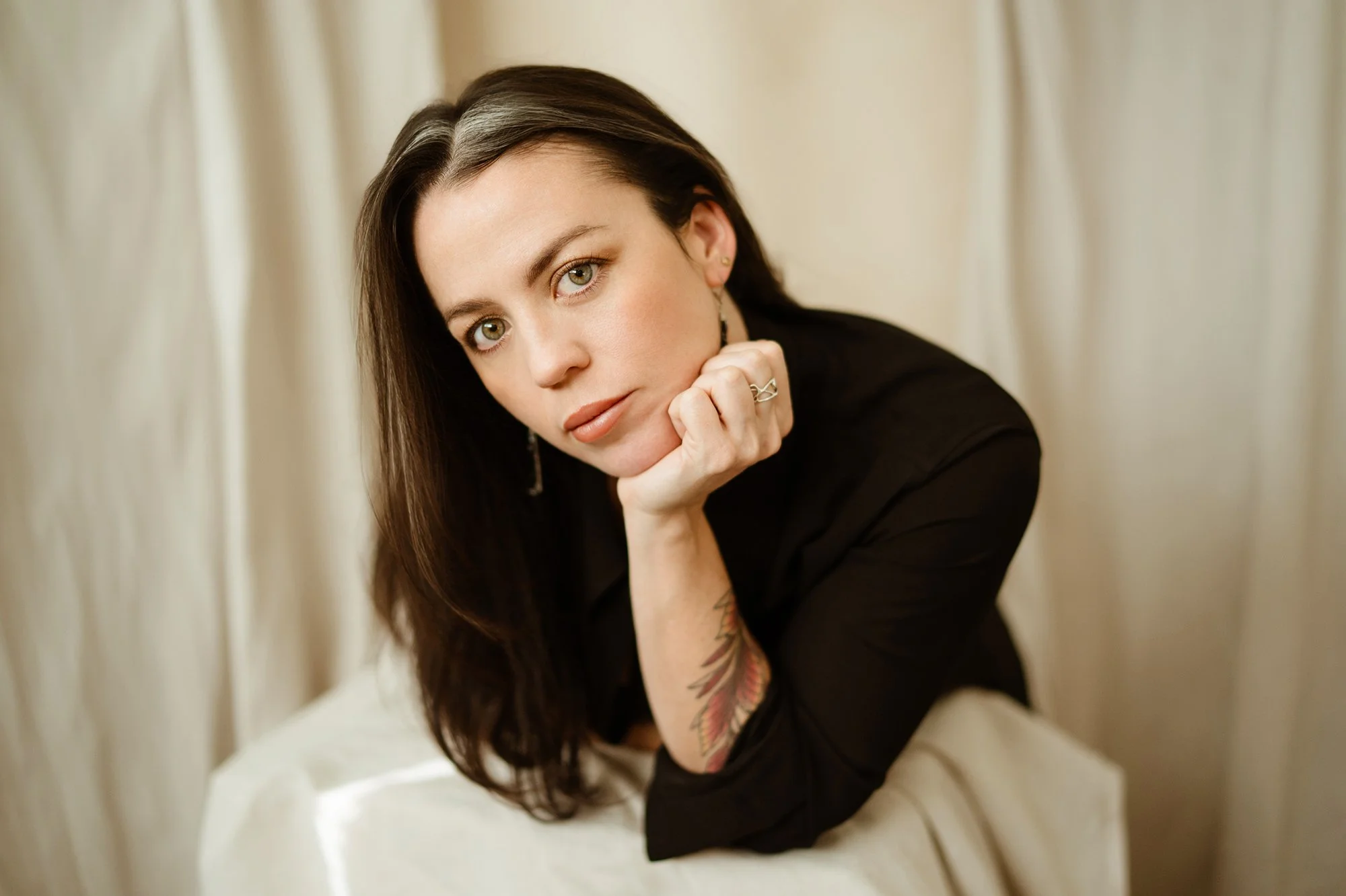 A woman with dark hair, wearing earrings and a black top, resting her chin on her hand, looking directly at the camera, with a neutral background.