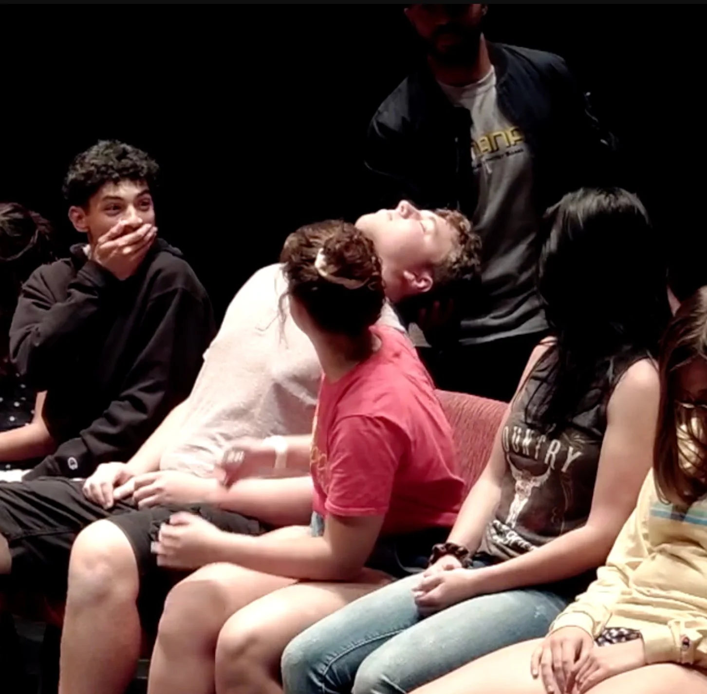 Group of young people sitting in a theater, reacting with laughter and surprise. One person in black covering mouth, others smiling or looking amused, in a dark setting.