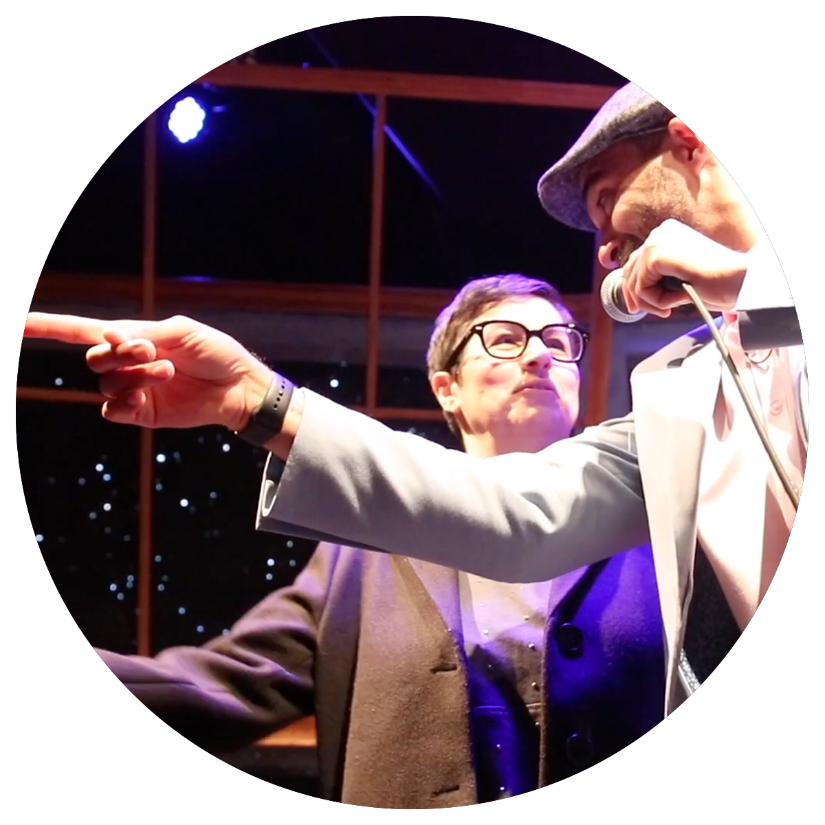 Two men are on stage, one with a microphone wearing a hat and the other wearing glasses and a blazer, with the man in the blazer pointing at something.