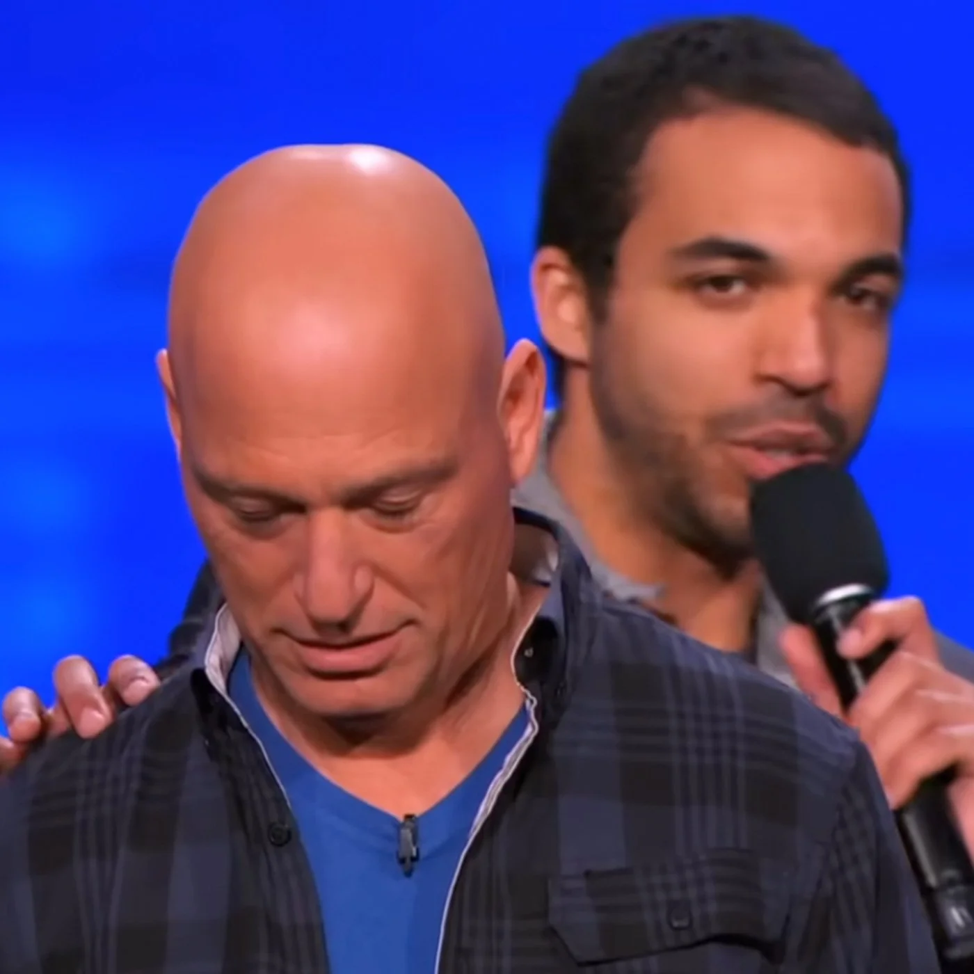 Two men on stage, one bald and the other with dark hair, scene from a game show or talk show with a blue background. The man with dark hair is speaking into a microphone and the bald man is looking down.