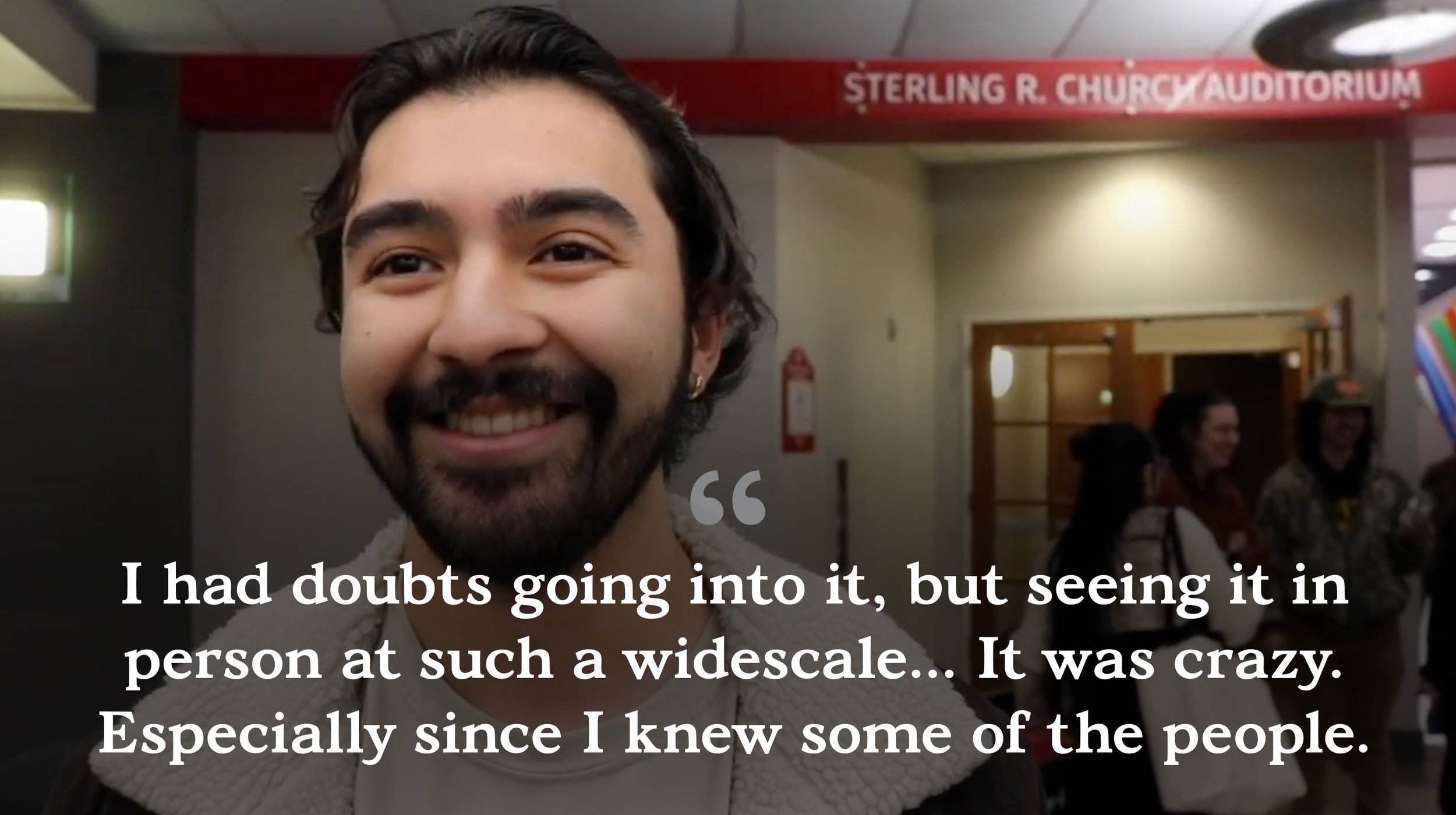 Smiling man with dark hair and a beard stands in an indoor space, with a red sign reading 'Sterling R. Church Auditorium' in the background. There are other people nearby, some of whom are also smiling.
