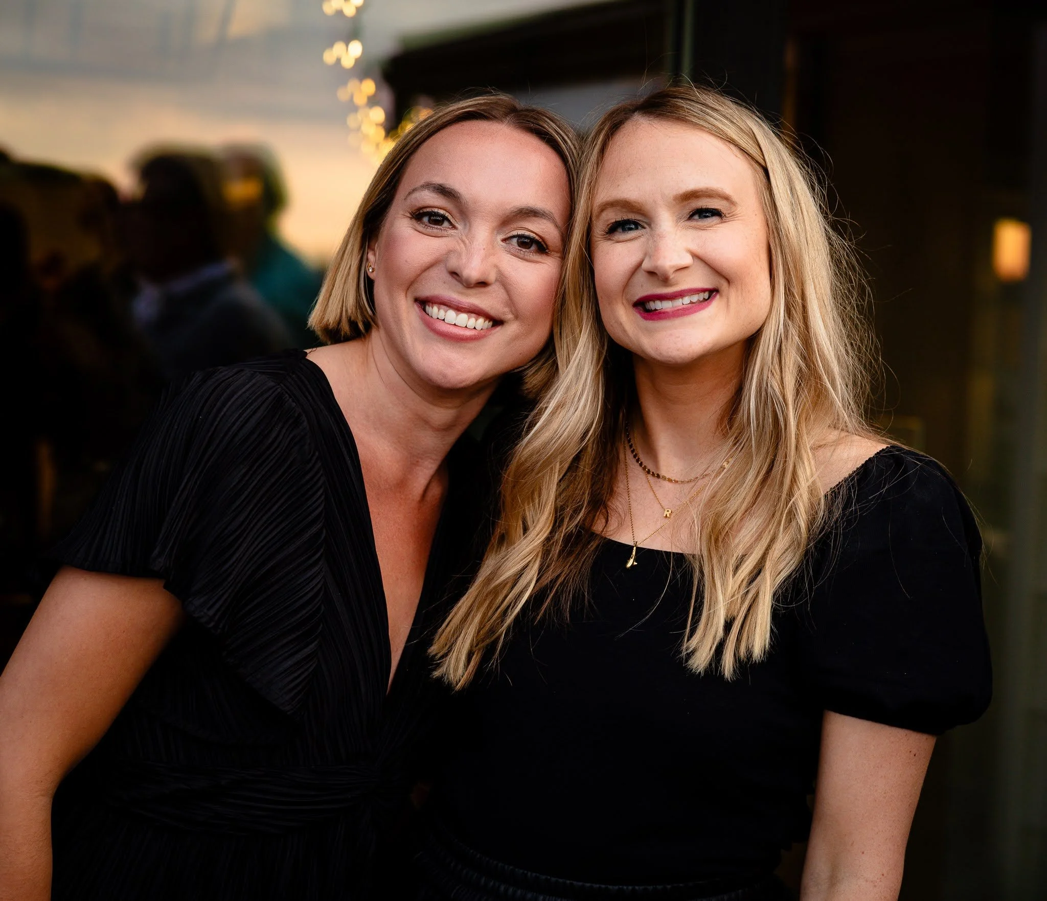 Two women smiling and posing together at an evening event, with blurred background and string lights.