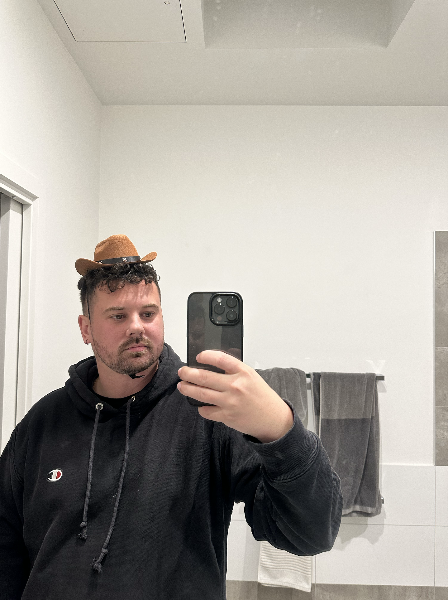 Codee Mac taking a selfie in a bathroom mirror. He is wearing a black hoodie and a small brown felt cowboy hat. The bathroom has a towel hanging on a bar and a white wall.