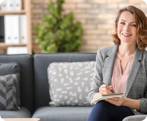 A woman with shoulder-length brown hair, wearing a gray plaid blazer and pink blouse, sitting on a couch with a notebook and pen, smiling in a cozy living room with a brick wall, bookshelves, and a green plant in the background.