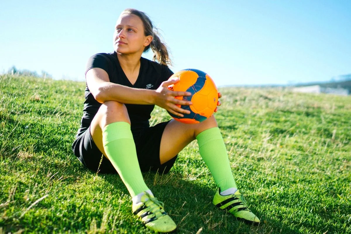 A female soccer playing holding a soccer ball sitting in the grass