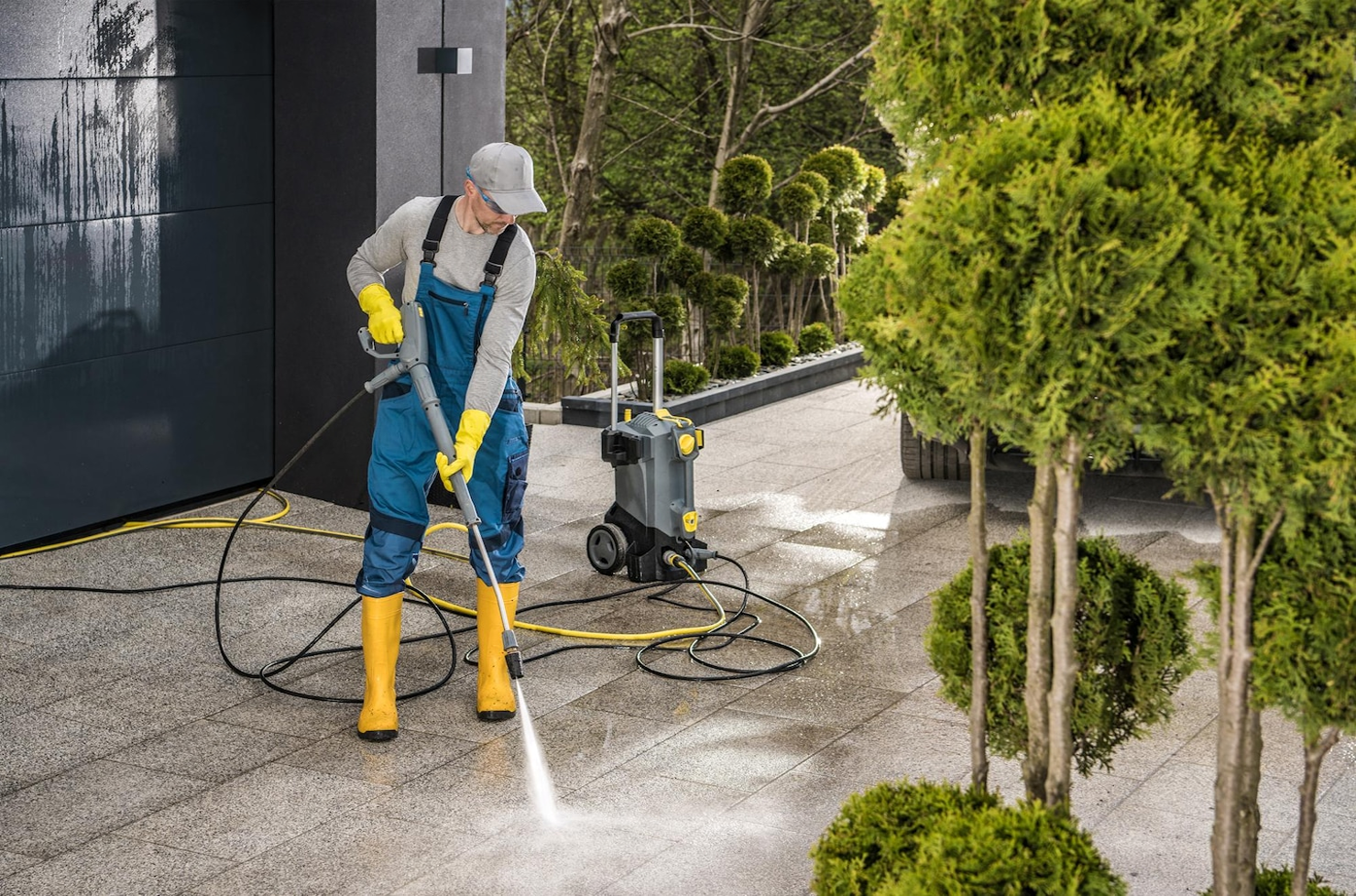 A worker wearing a gray cap, gray shirt, blue overalls, yellow gloves, and yellow waterproof boots is cleaning an outdoor tiled surface with a pressure washer. There is a black building wall to the left and a row of trimmed green bushes to the right. The worker is focused on cleaning the tiles, and a portable pressure washer is connected to the pressure washer wand. The scene appears to be in a well-maintained outdoor area.