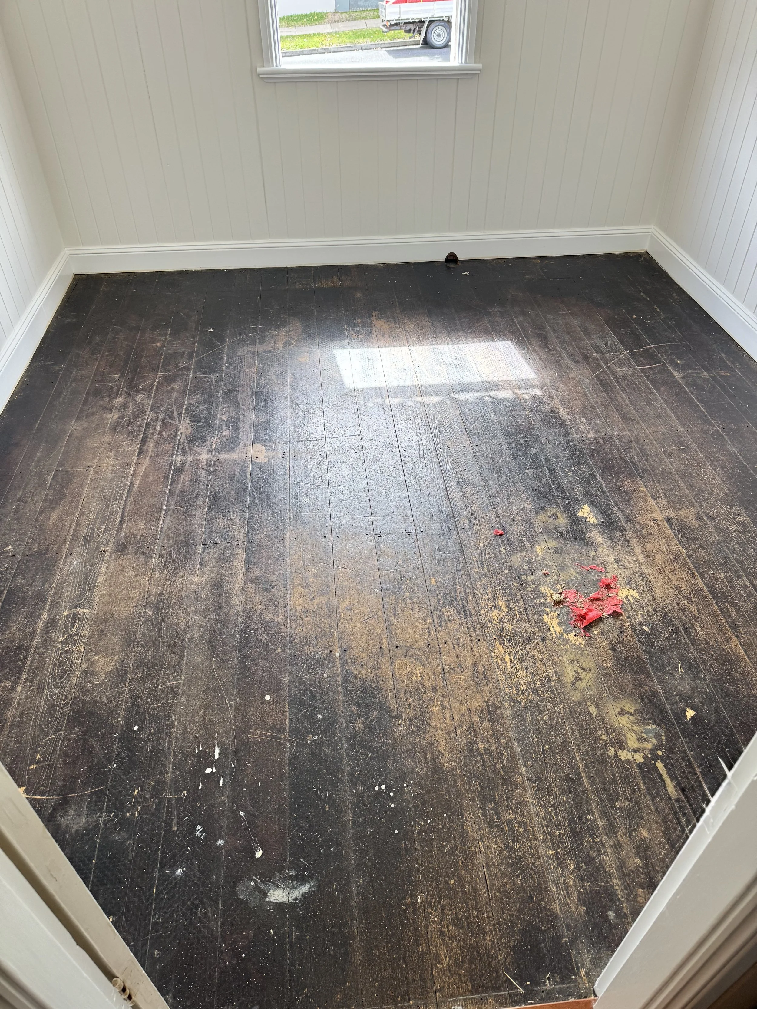 Empty room with dark wooden floor, dirt and debris, and a small patch of red-painted wood debris on the floor. The room has cream-white paneled walls and a window showing a street view outside.