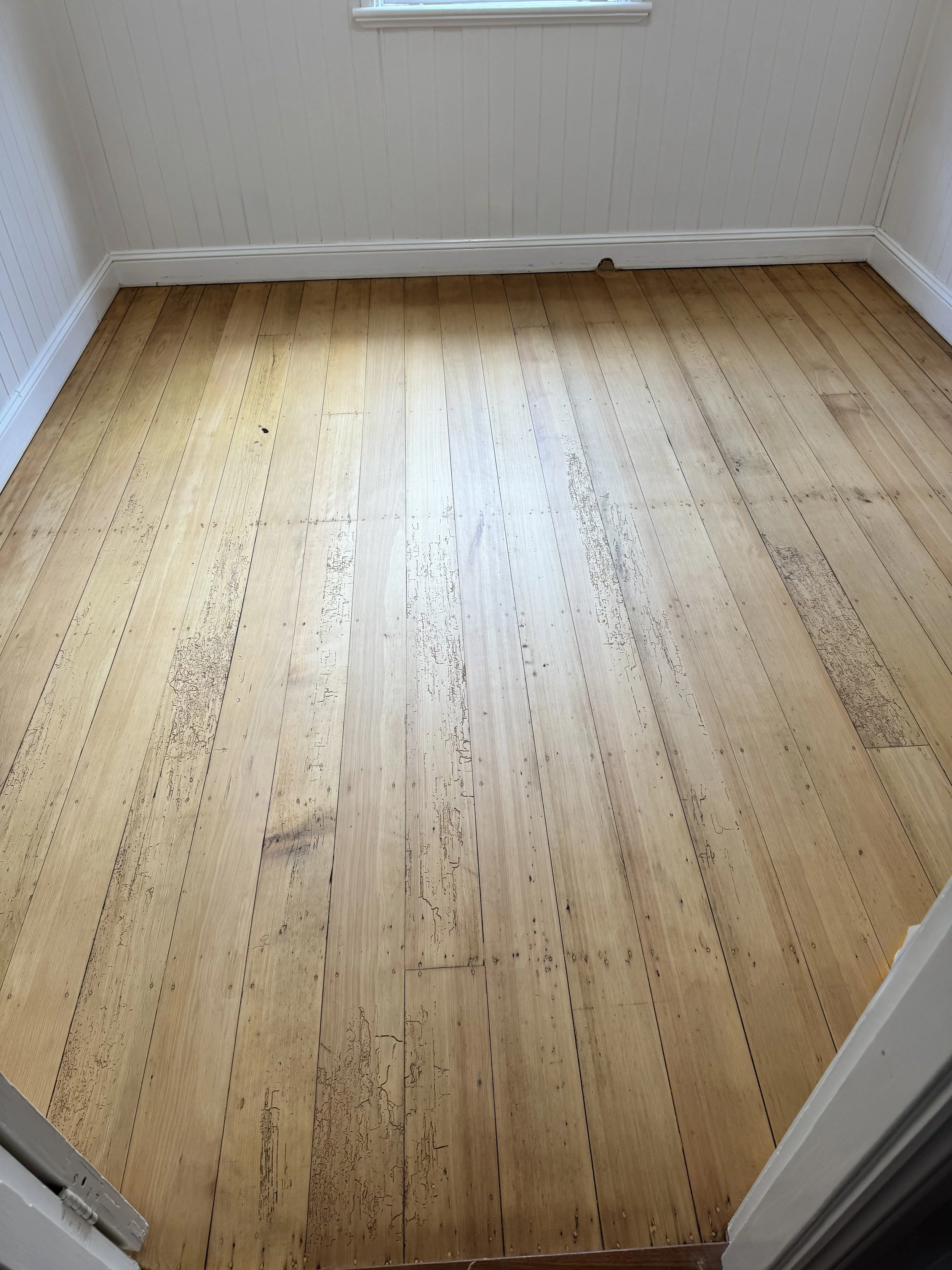 Room with hardwood floor and white-paneled walls, some wear and scratches on the flooring, with a white baseboard and partially visible window.