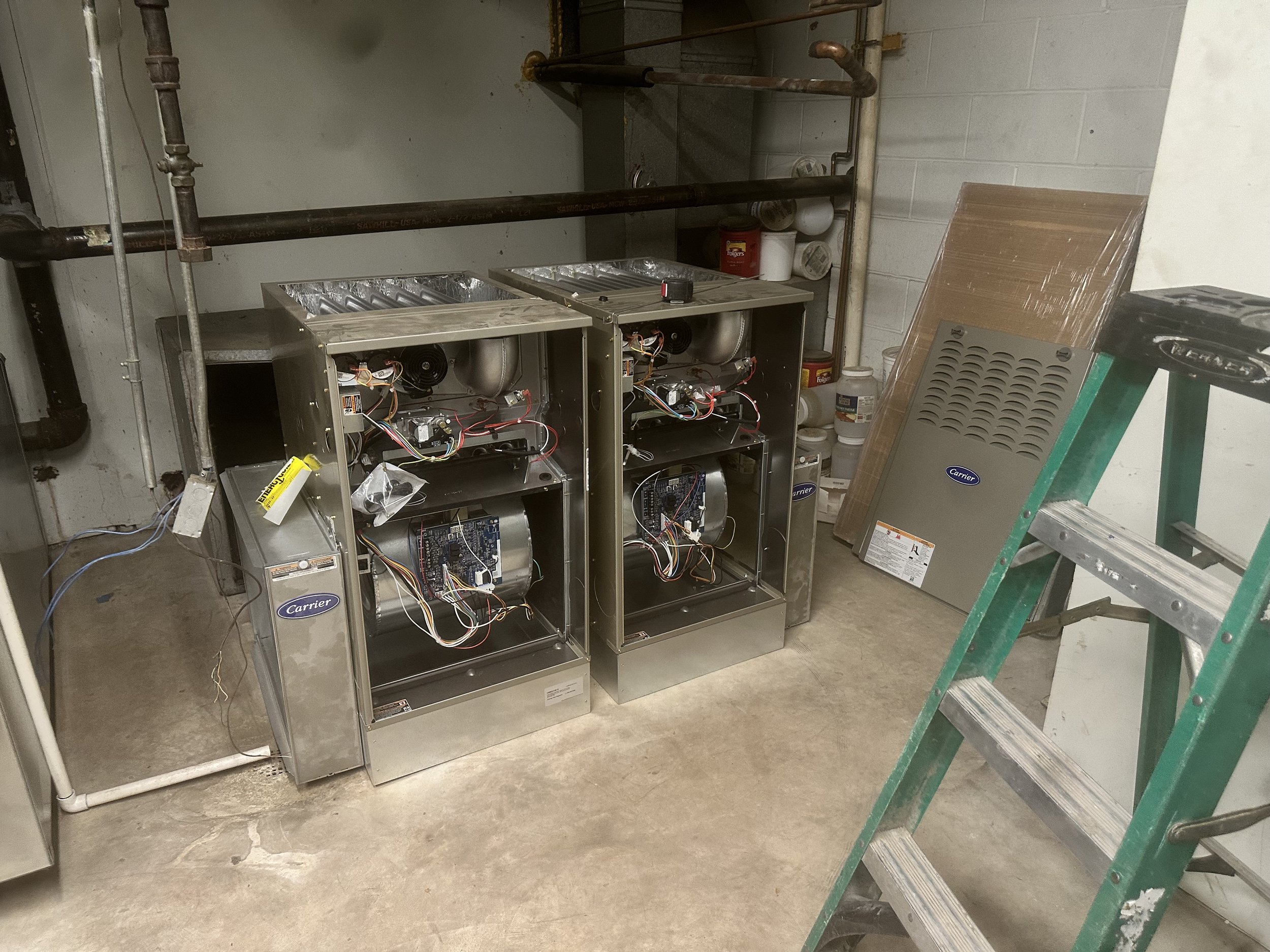 Two air conditioning units with open covers and visible internal components, placed on a concrete floor in a utility room with pipes on the wall.