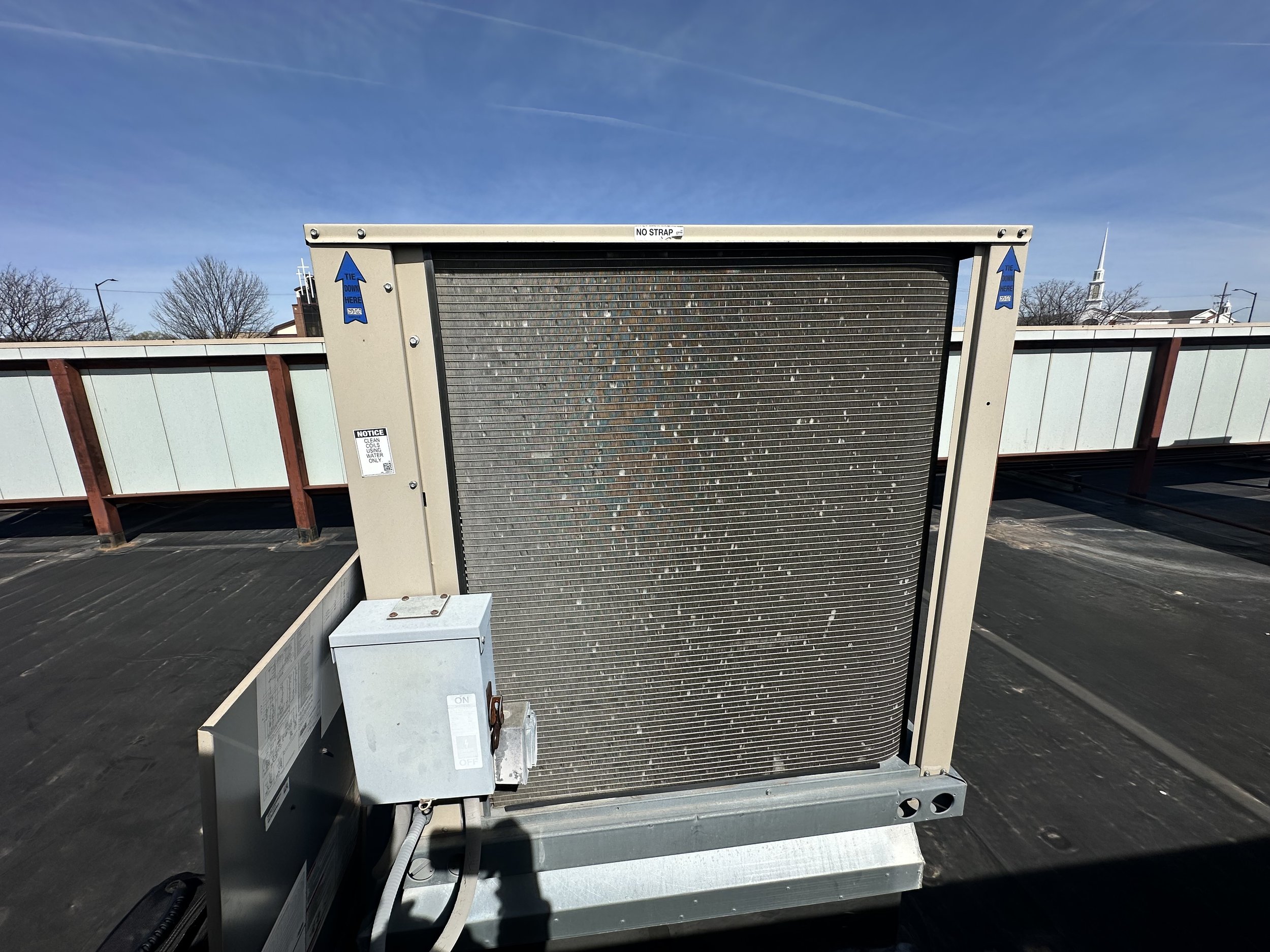 Rooftop HVAC unit with a large metal grille, electrical box, and rooftop safety railing, under a blue sky.