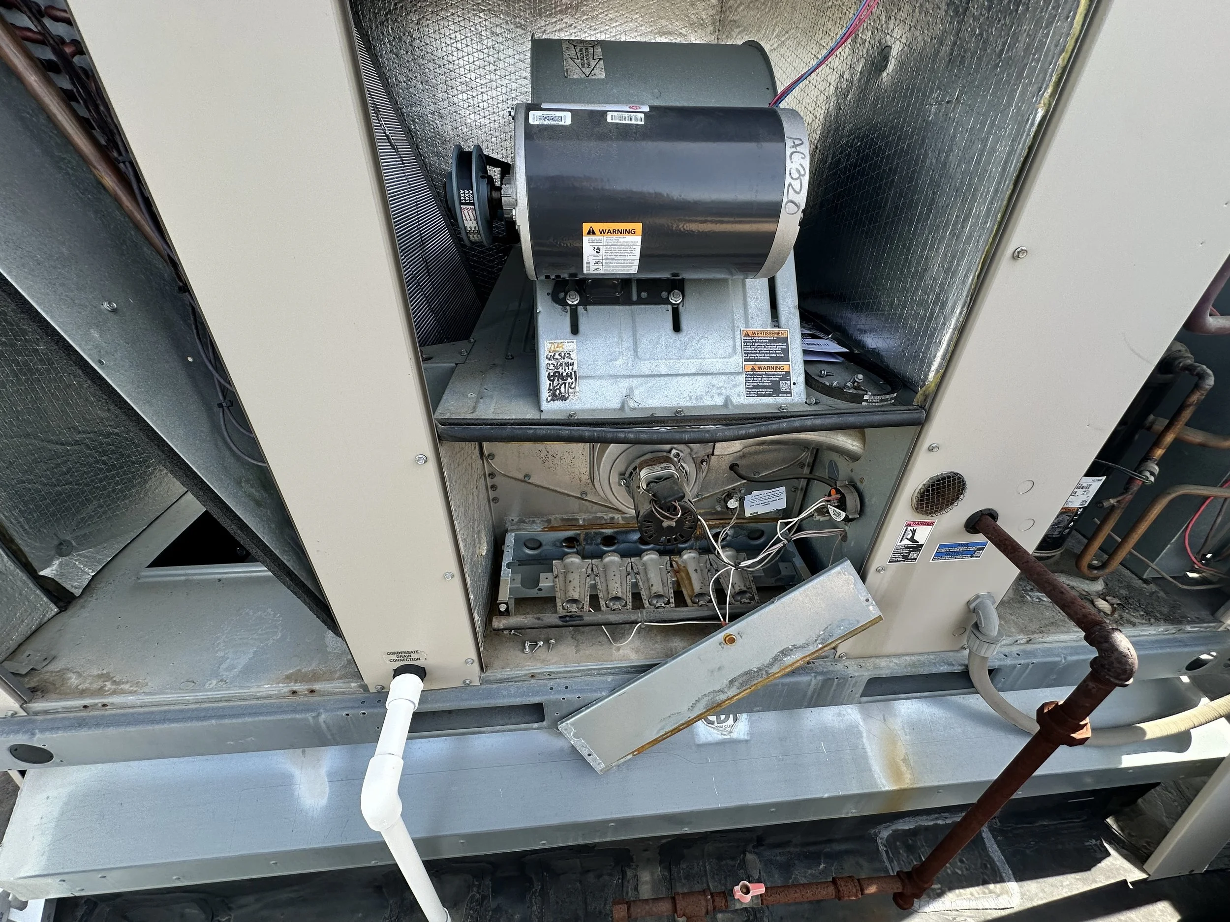 Inside view of an HVAC unit's interior with some components exposed, including a black motor, metal panels, wiring, and refrigerant lines, with some rust on pipes and a white PVC pipe at the bottom.