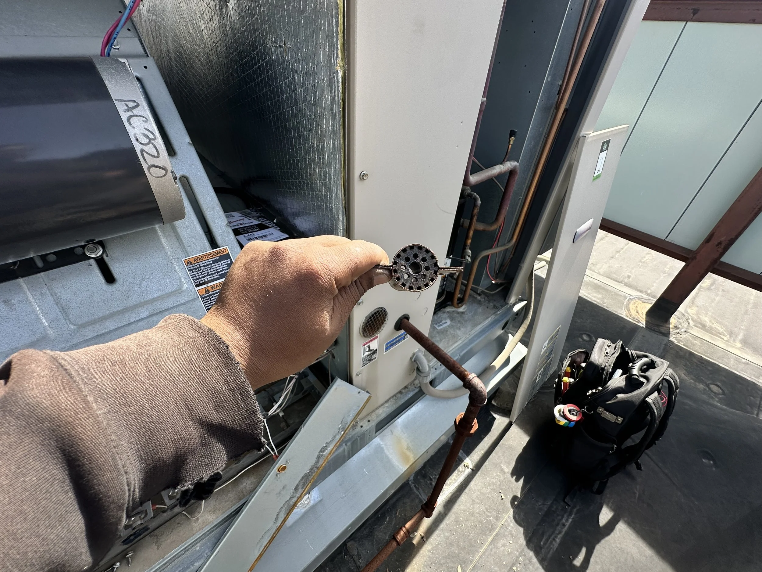 A technician's hand holding a refrigerant valve service port on an outdoor air conditioning unit, with the cover removed showing the internal copper pipes and components, on a rooftop or outdoor area.
