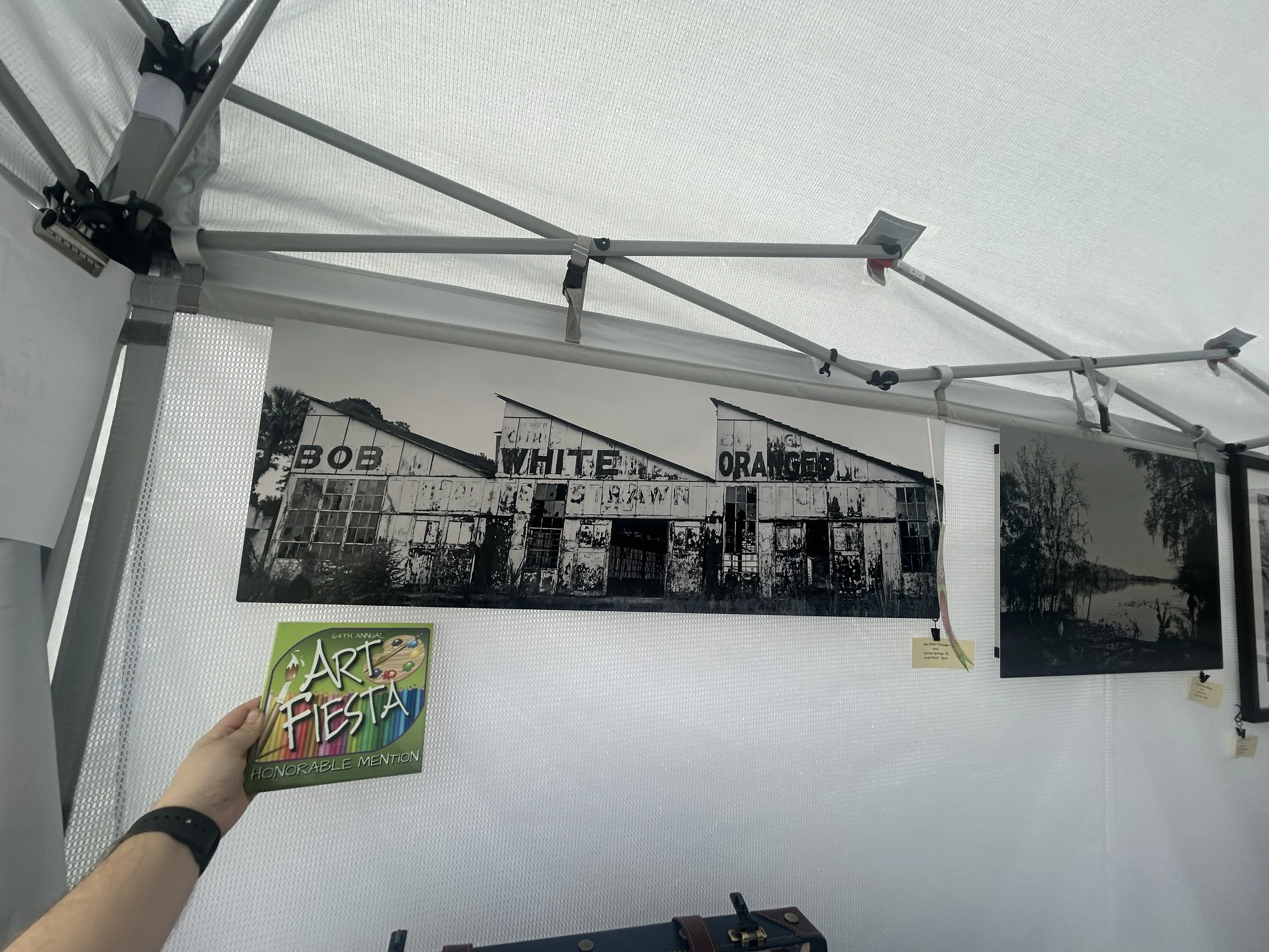 A black and white photo of an old barn with signs reading 'BOB', 'WHITE', 'ORANGES'. A hand is holding an art award plaque that says 'ART FIESTA HONORABLE MENTION'.