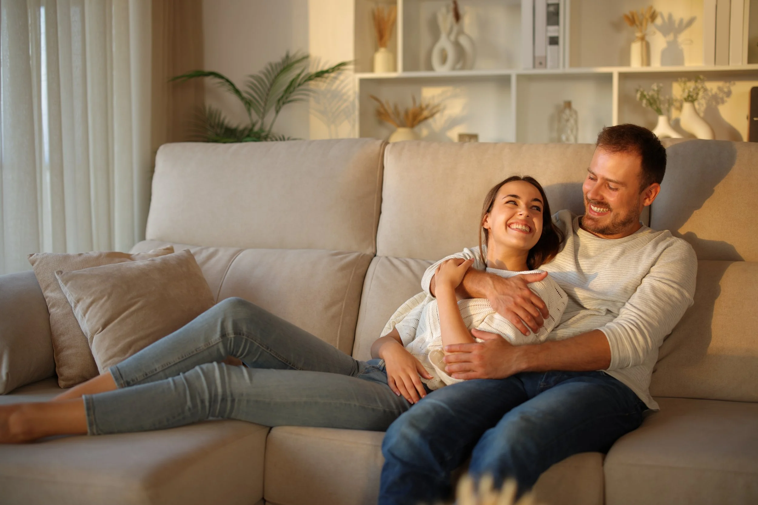 A man and woman sitting on a beige sofa, smiling and embracing each other, in a living room with decorative shelves, vases, and a large green potted plant.
