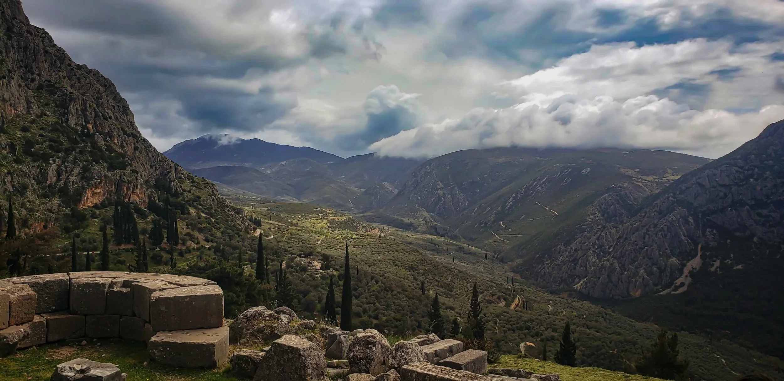 A view of a mountain valley with green hills, rocky slopes, scattered trees, and cloudy sky.