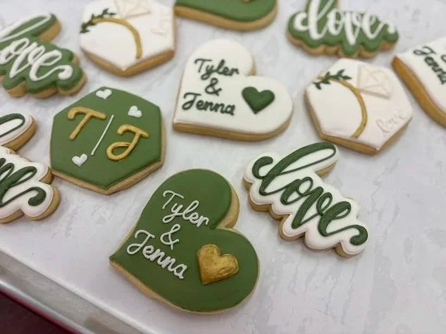 Decorated cookies with love and couple themes, featuring names Tyler and Jenna, hearts, and wedding rings in green, white, and gold colors.