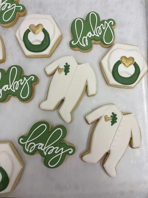 Decorative cookies with Christmas themes, including the words "baby" and "galaxy" in green script, and white baby onesies with gold hearts and green shamrocks.