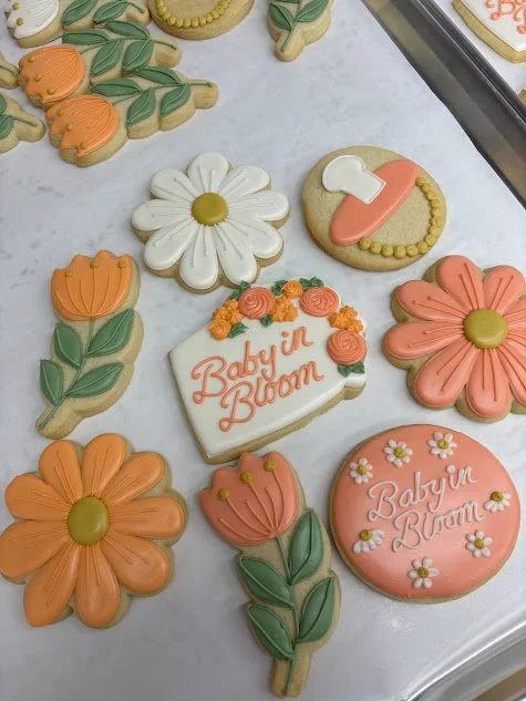 Decorated cookies with flowers, a baby shoe, and potted plant motifs, including the words "Baby in Bloom" on a rectangular cookie and a round cookie.