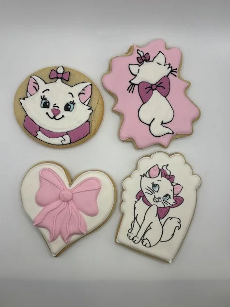 Four decorated cookies with a cat theme. Top left cookie features a smiling white cat with a pink bow and a purple collar. Top right cookie shows a white cat with a purple bow on a pink background, rubbing its head. Bottom left cookie is a pink bow o