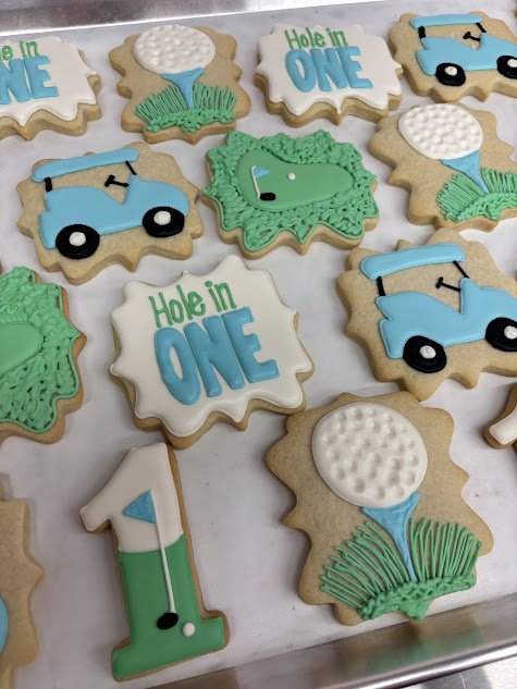 Cookies decorated for a golf-themed celebration, including golf balls, golf carts, and signs that say "Hole in ONE."