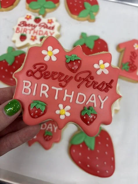 Decorated cookie with pink icing, strawberries, white daisies, and green leaves, reading "Berry First BIRTHDAY."