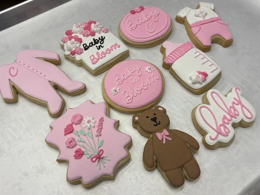 Cookies with pink and white icing, decorated with baby-themed designs including a teddy bear, flowers, and messages like 'Baby in Bloom' and 'Baby'. Conference table surface with cookies displayed.