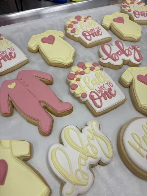 Decorated sugar cookies with baby shower themes, including ones shaped like baby onesies, a pink diaper, and text such as "You're Expecting," "Hatch," and "All Ways Wonder."