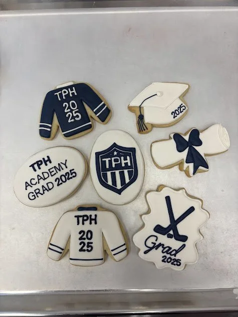 Assorted decorated cookies celebrating graduation in 2025, including a varsity jacket, a cap, a shield with "TPH" on it, a diploma, a bow, a sweater with "TPH ACADEMY GRAD 2025," and a sword with "Grad 2025" on it.