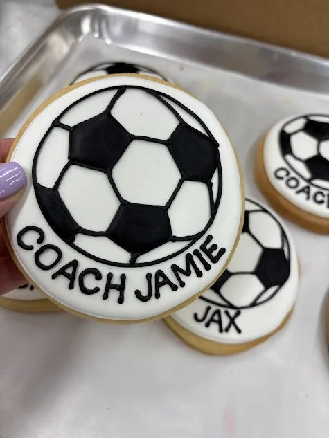 Decorative cookie with a soccer ball design and the words 'Coach Jamie' written on it.