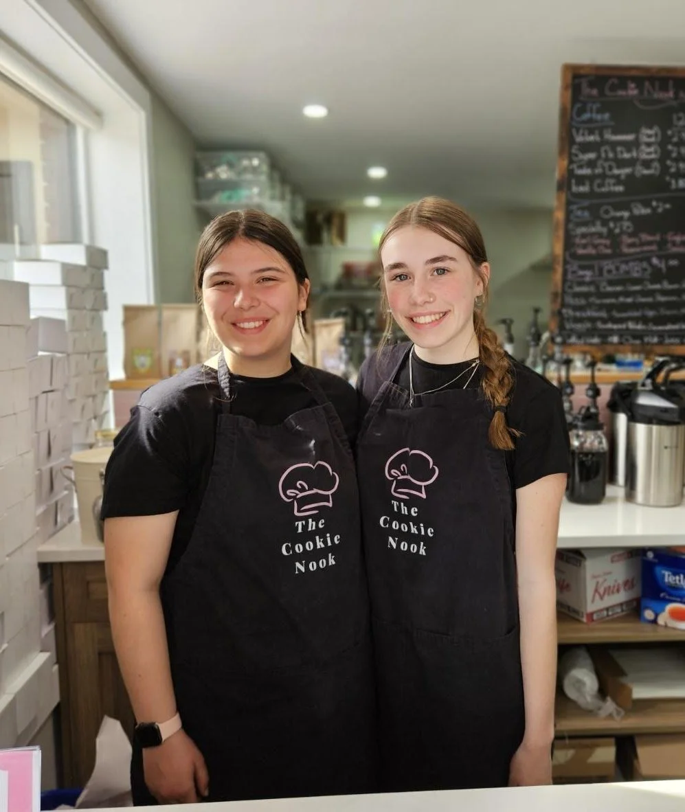 Two young women smiling in a bakery, wearing black aprons with a cookie chef logo that says 'The Cookie Nook', inside a café.