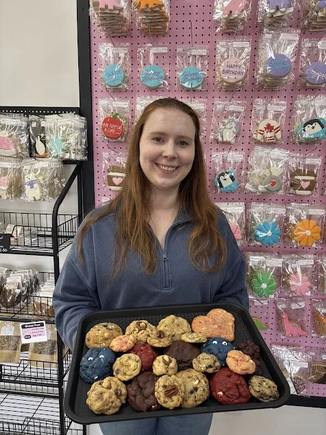 A young woman with long reddish hair smiling and holding a tray of assorted cookies in a store decorated with birthday-themed cookies on a pink display background.