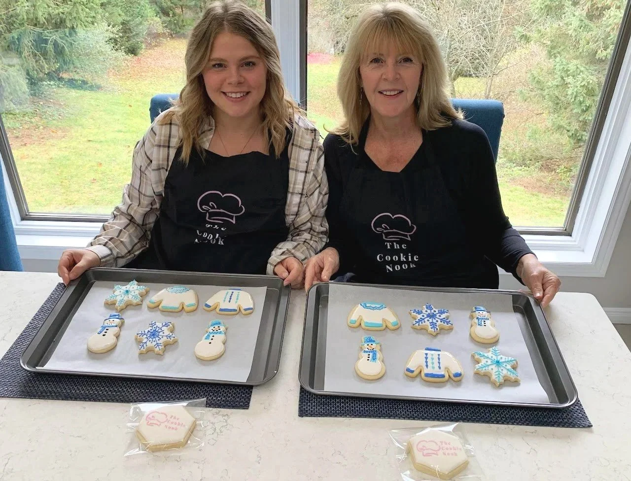 Two women sitting at a kitchen table holding cookie trays with decorated holiday cookies, including snowflakes, snowmen, Christmas sweaters, and mittens, in front of large windows showing greenery outside.