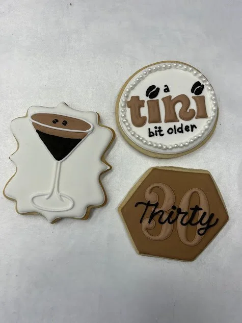 Three decorated cookies on a white surface: one with a martini glass and an olive, one round with the words "a tini bit older" and decorated with pearls and coffee beans, and one hexagonal with the number "30" and the word "thirty".